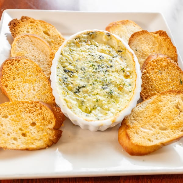Creamy spinach dip in a white oval dish, surrounded by toasted baguette slices on a white plate.