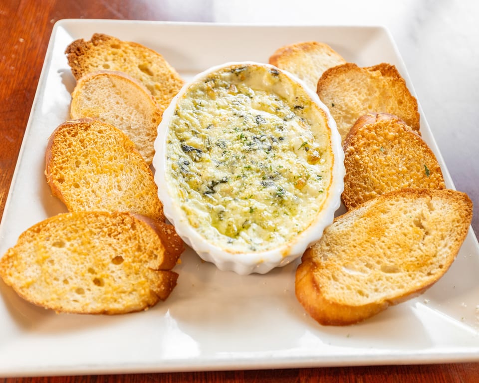 Creamy spinach dip in a white oval dish, surrounded by toasted baguette slices on a white plate.