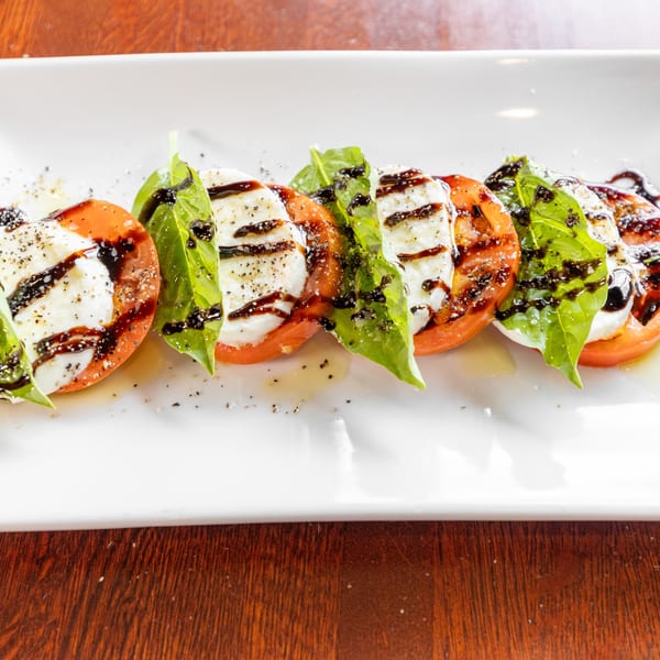 Caprese salad with sliced tomatoes, mozzarella, basil, balsamic glaze, and pepper on a white plate.