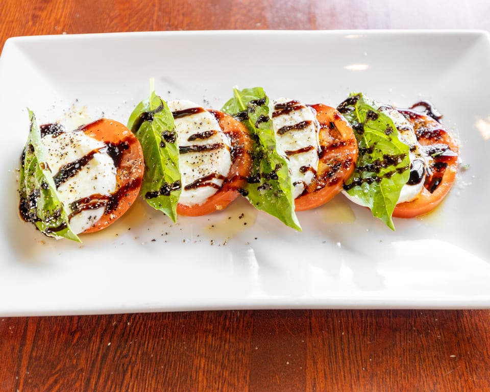 Caprese salad with sliced tomatoes, mozzarella, basil, balsamic glaze, and pepper on a white plate.