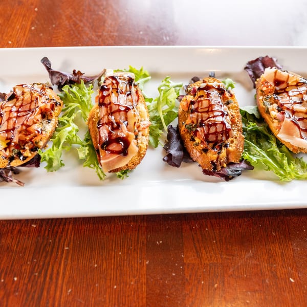 Four bruschetta appetizers with prosciutto, cheese, and balsamic glaze on a white plate with lettuce.