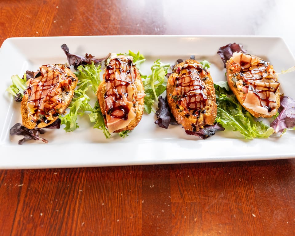 Four bruschetta appetizers with prosciutto, cheese, and balsamic glaze on a white plate with lettuce.