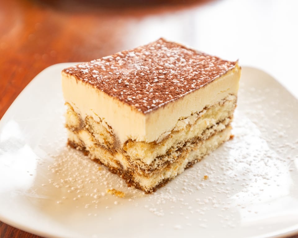 A square slice of tiramisu with layers of cake and cream, dusted with cocoa and powdered sugar.