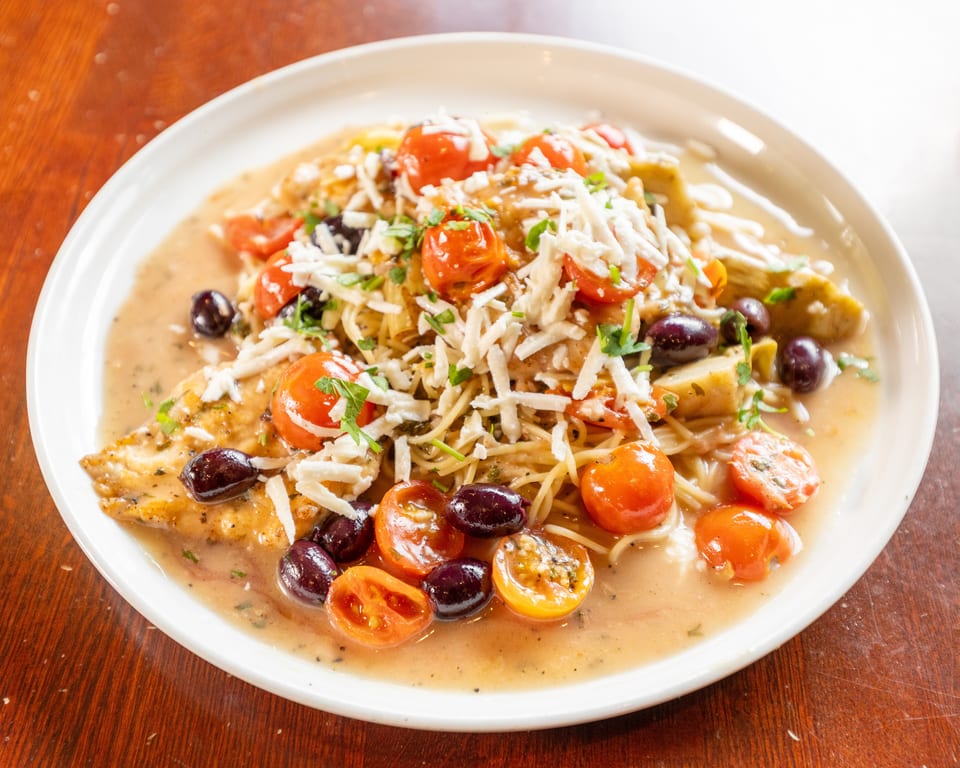 Chicken piccata with spaghetti, cherry tomatoes, olives, and shredded cheese in a creamy sauce.
