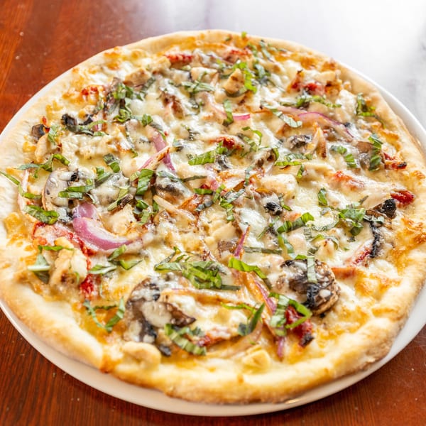 A whole round pizza with chicken, mushrooms, red onions, and basil on a white plate.