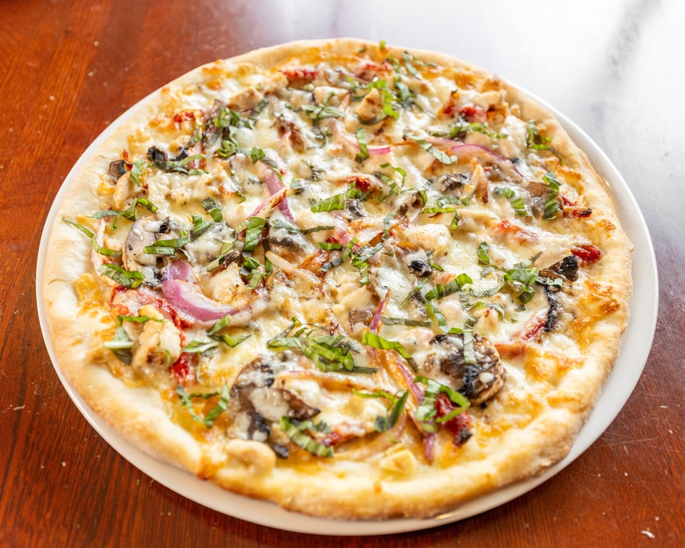 A whole round pizza with chicken, mushrooms, red onions, and basil on a white plate.
