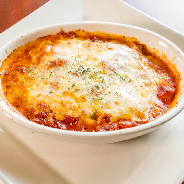 Baked pasta dish with melted cheese and red sauce, garnished with herbs, in a white oval bowl.