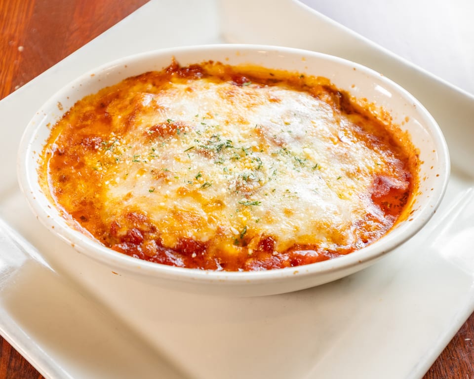 Baked pasta dish with melted cheese and red sauce, garnished with herbs, in a white oval bowl.