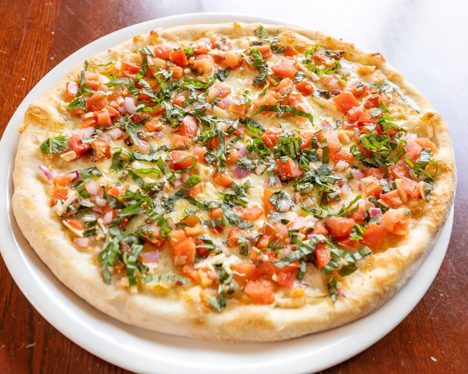 A round bruschetta pizza with diced tomatoes, red onions, and fresh basil on a white plate.
