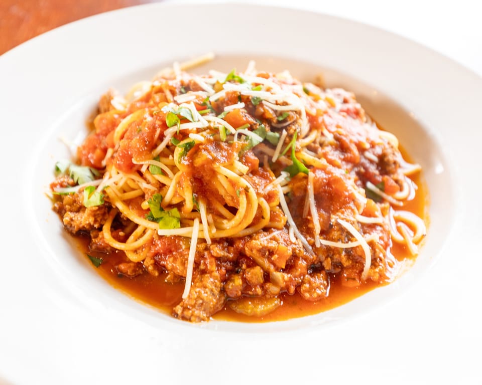 Spaghetti with meat sauce, parmesan cheese, and fresh herbs in a white bowl.