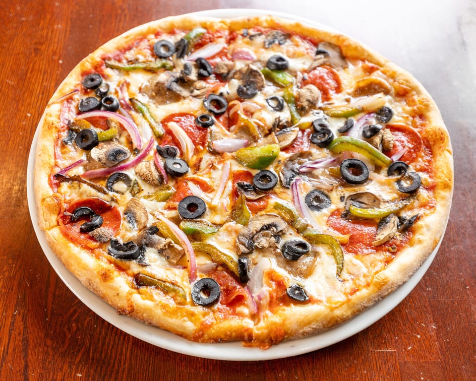A whole pizza with pepperoni, mushrooms, olives, green peppers, and red onions on a white plate.