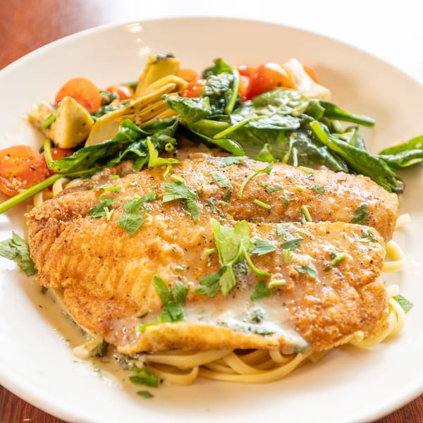 Fried fish fillets with creamy sauce, pasta, spinach, cherry tomatoes, and artichoke hearts on a white plate.