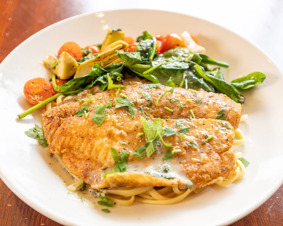 Fried fish fillets with creamy sauce, pasta, spinach, cherry tomatoes, and artichoke hearts on a white plate.