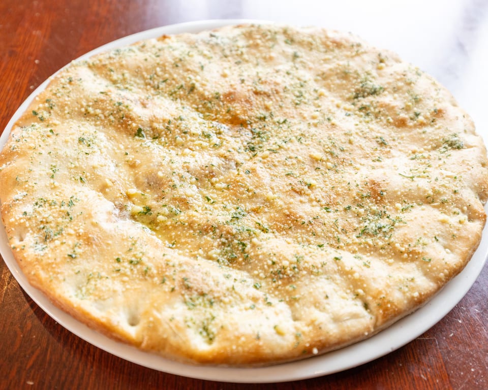 A round, golden-brown garlic bread with melted cheese and green herbs on a white plate.