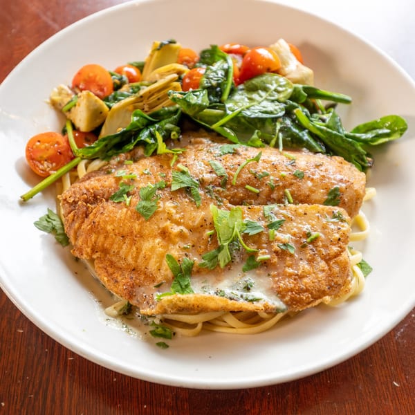 Fried fish fillets with pasta, spinach, cherry tomatoes, and artichoke hearts on a white plate.