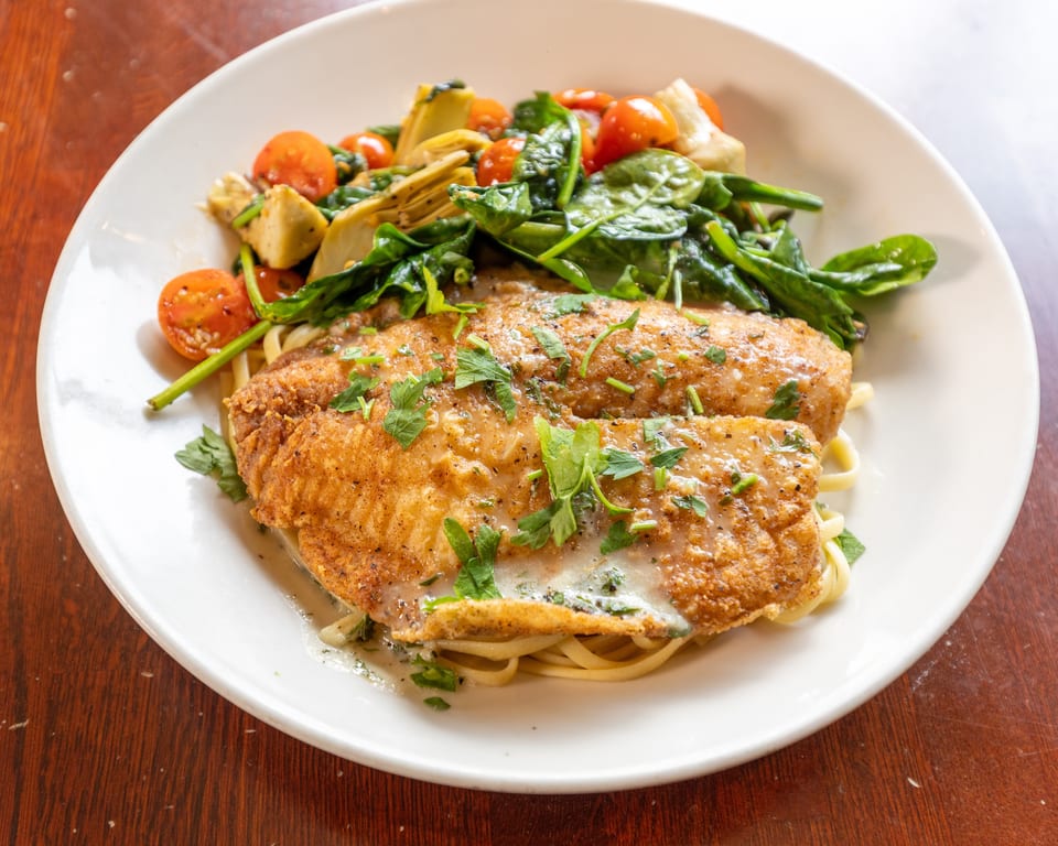 Fried fish fillets with pasta, spinach, cherry tomatoes, and artichoke hearts on a white plate.