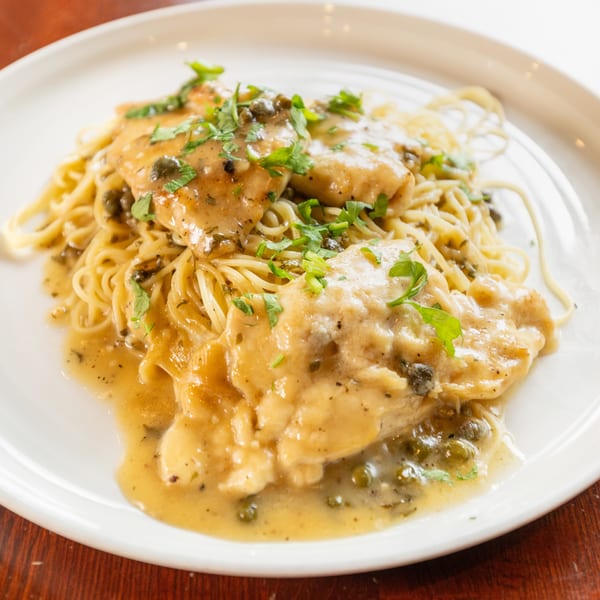 Chicken piccata with capers and parsley over spaghetti on a white plate.