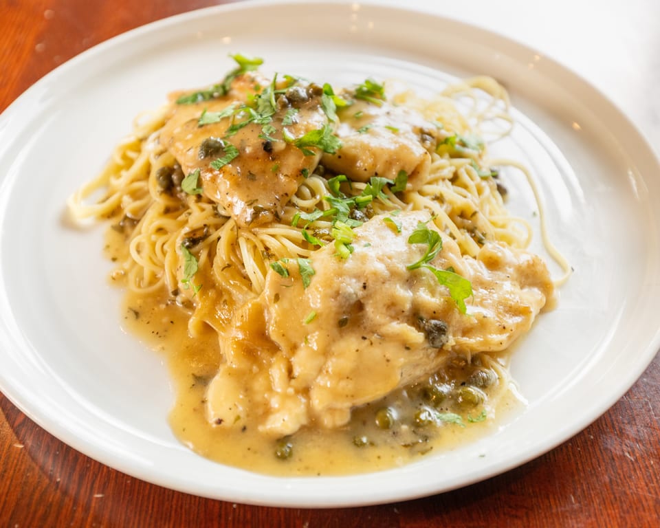 Chicken piccata with capers and parsley over spaghetti on a white plate.