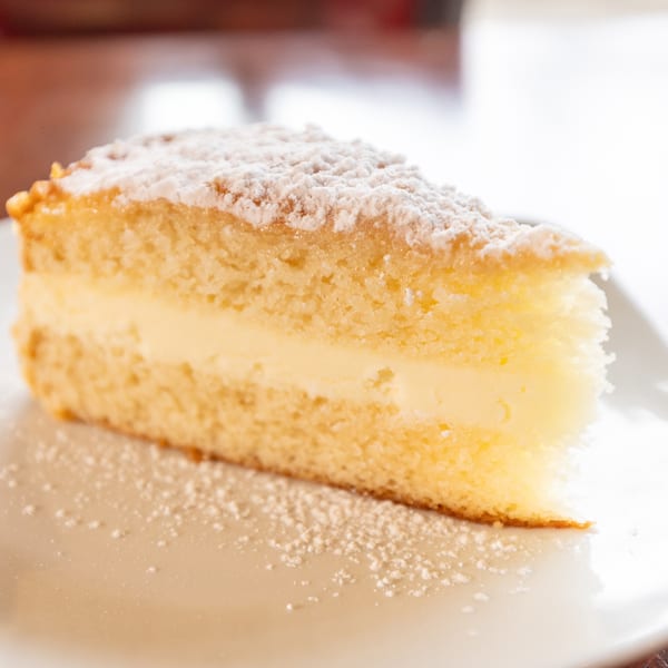 A slice of vanilla cake with cream filling and powdered sugar on a white plate.