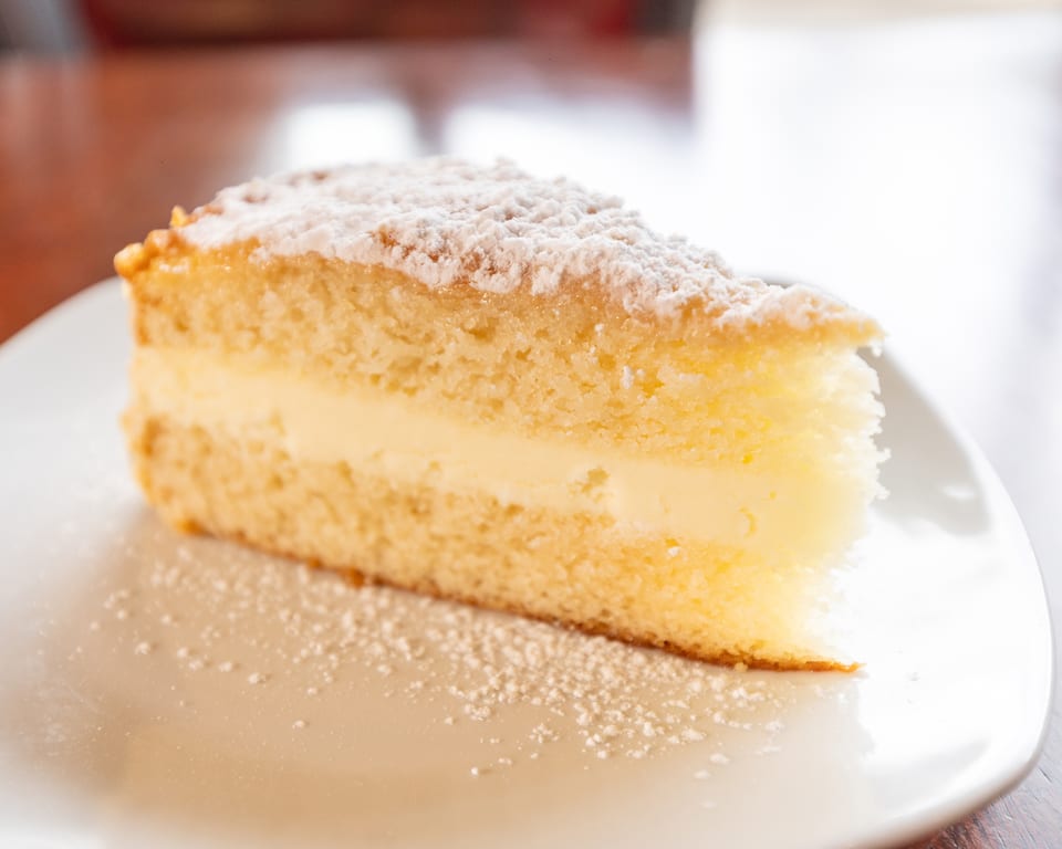 A slice of vanilla cake with cream filling and powdered sugar on a white plate.