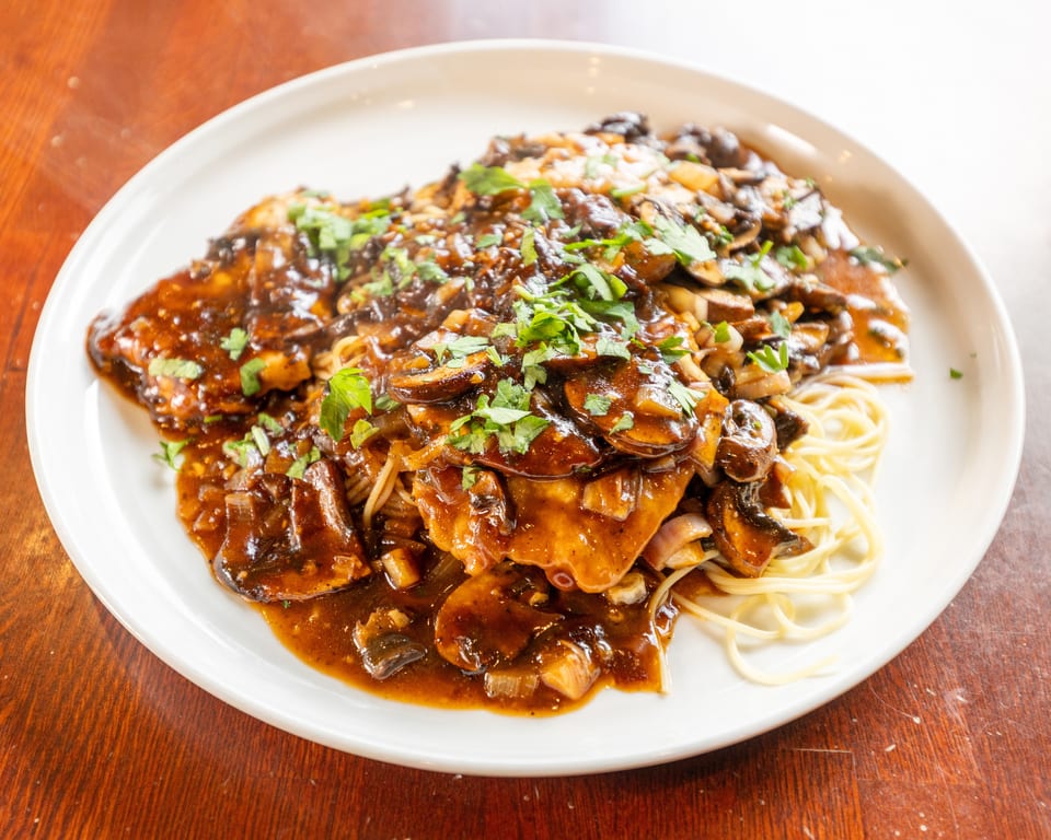 Chicken Marsala with mushrooms, onions, and parsley over spaghetti on a white plate.