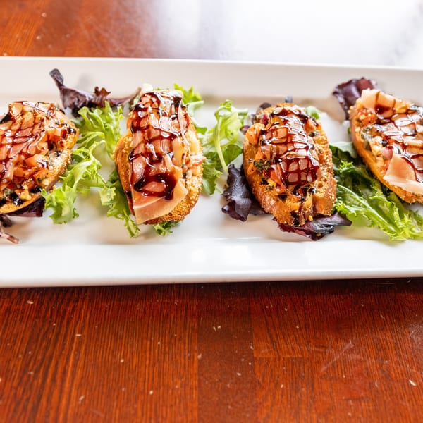 Four bruschetta appetizers with prosciutto, diced tomatoes, and balsamic glaze on a white platter.