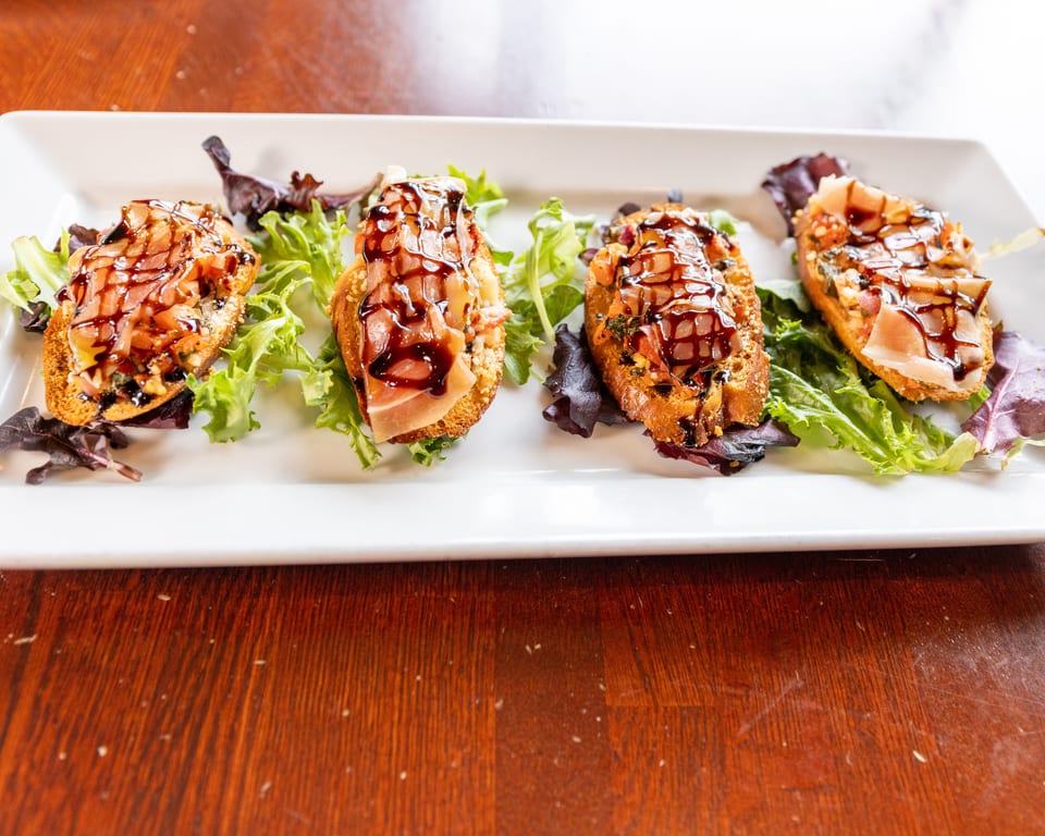 Four bruschetta appetizers with prosciutto, diced tomatoes, and balsamic glaze on a white platter.
