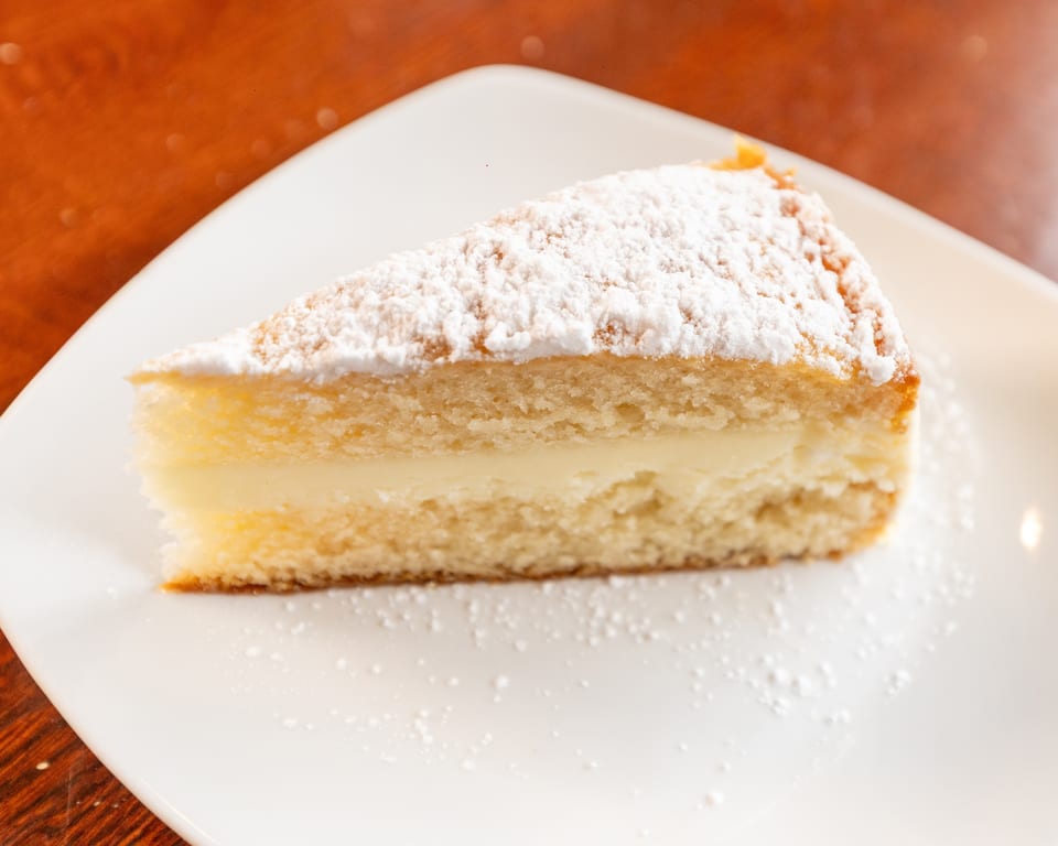 A slice of vanilla cake with a creamy filling and powdered sugar on a white plate.