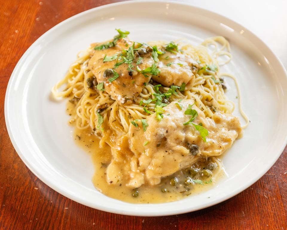 Chicken piccata with capers and parsley over spaghetti on a white plate.