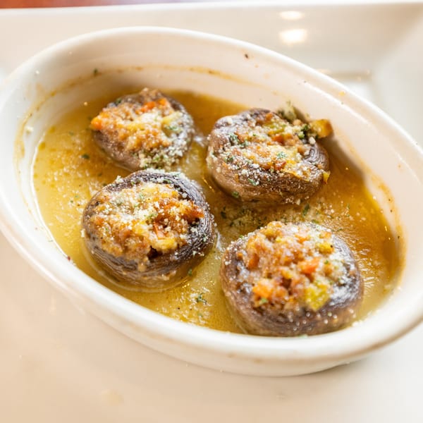 Four stuffed mushrooms in a white oval dish, swimming in a buttery sauce, topped with cheese and herbs.