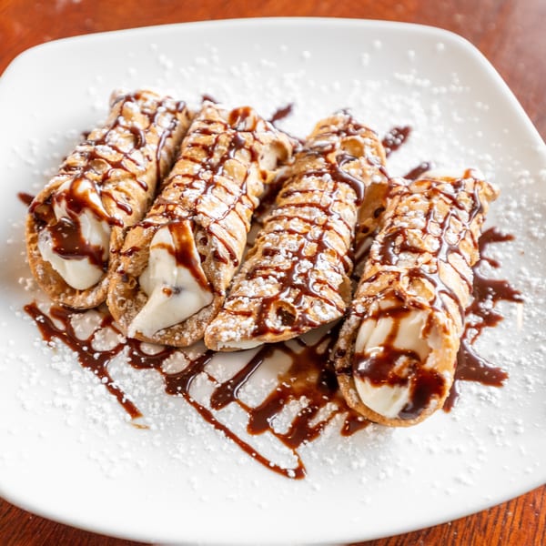 Four cannoli on a white plate, drizzled with chocolate syrup and powdered sugar.