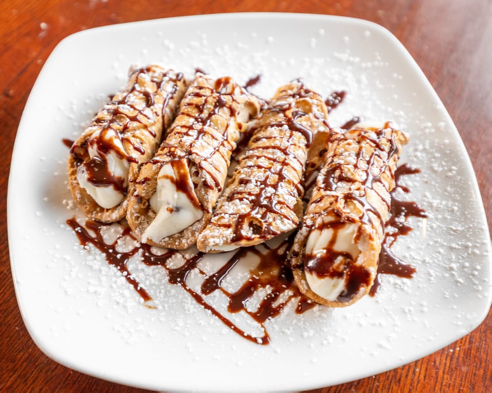 Four cannolis filled with cream, drizzled with chocolate sauce and powdered sugar on a white plate.