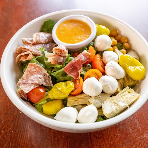 A vibrant Italian salad with salami, prosciutto, mozzarella, tomatoes, artichokes, and dressing in a white bowl.