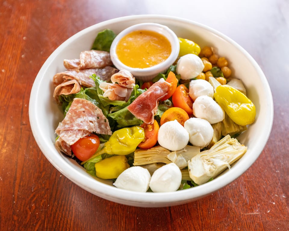 A vibrant Italian salad with salami, prosciutto, mozzarella, tomatoes, artichokes, and dressing in a white bowl.