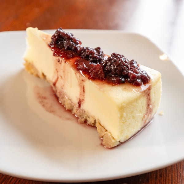 A slice of cheesecake with a graham cracker crust, topped with dark berry compote and syrup.