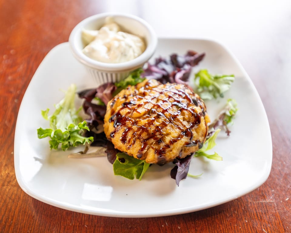 Golden-brown crab cake drizzled with balsamic glaze, served with mixed greens and a side of tartar sauce.