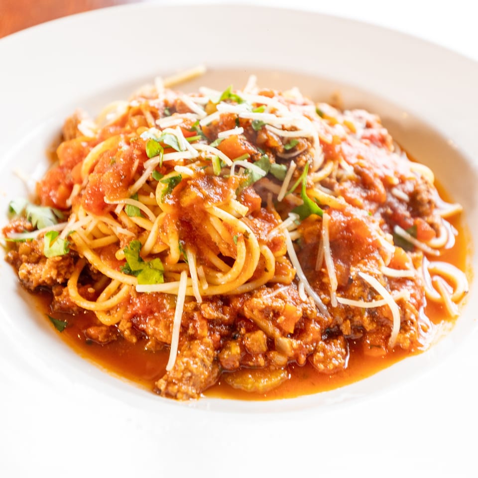 Spaghetti with meat sauce, parmesan cheese, and fresh herbs in a white bowl.