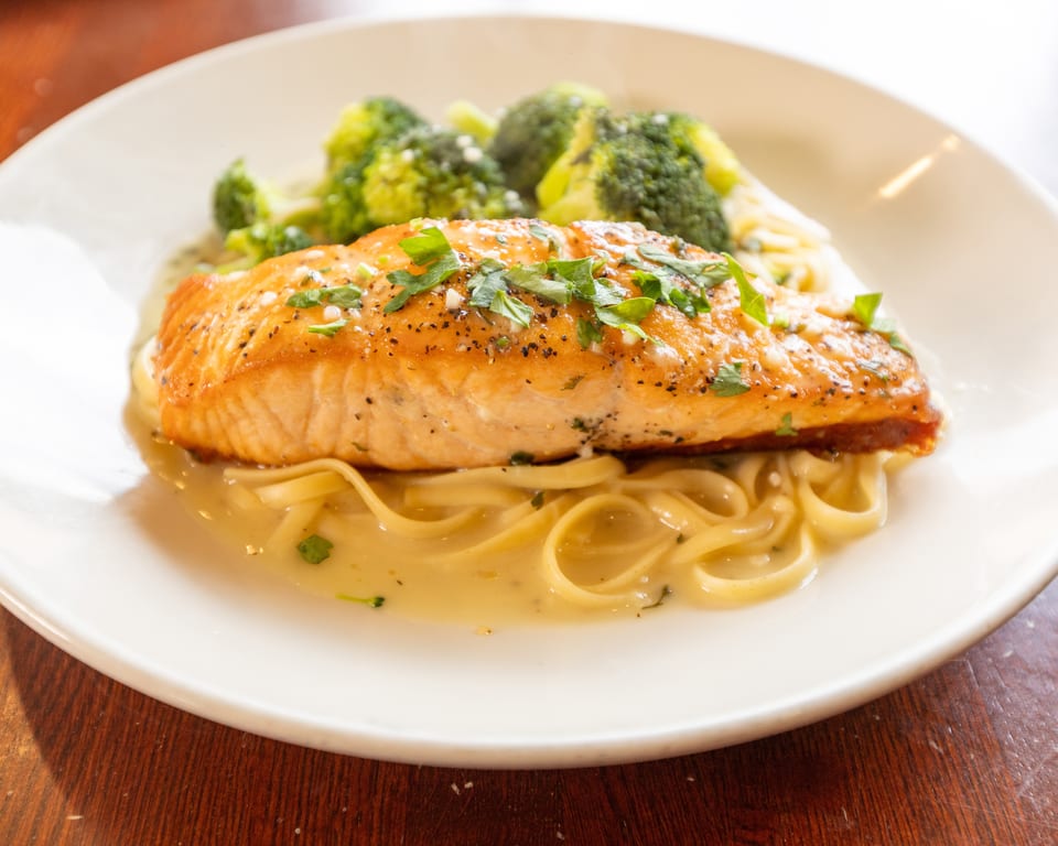 Pan-seared salmon with fresh parsley, linguine pasta in cream sauce, and steamed broccoli on a white plate.