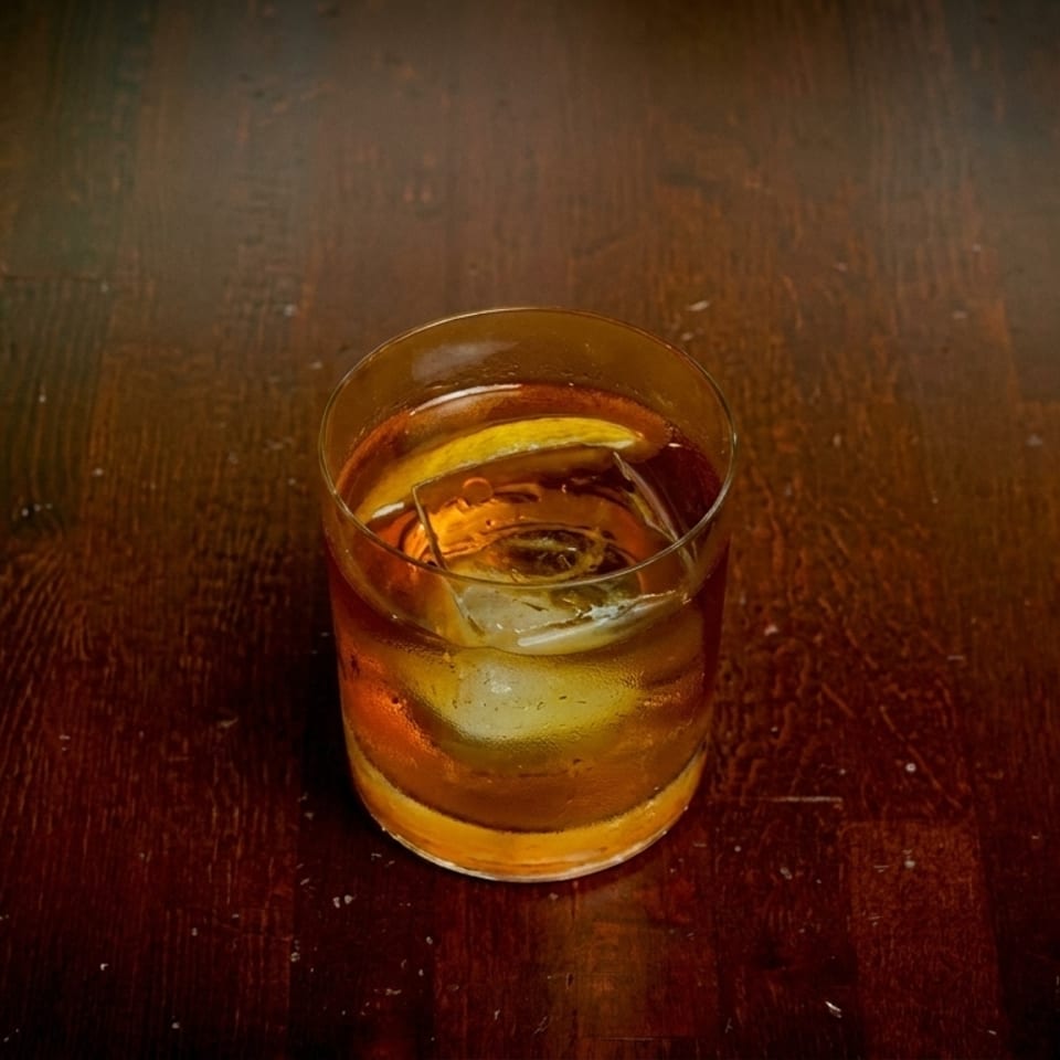 An amber cocktail with a large ice cube and citrus peel sits on a dark wooden table.