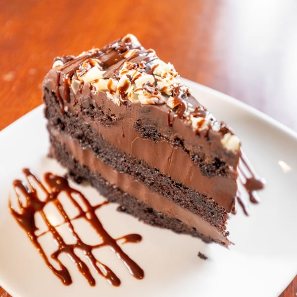 A slice of rich chocolate cake with chocolate frosting, drizzled with chocolate sauce and white chocolate shavings.