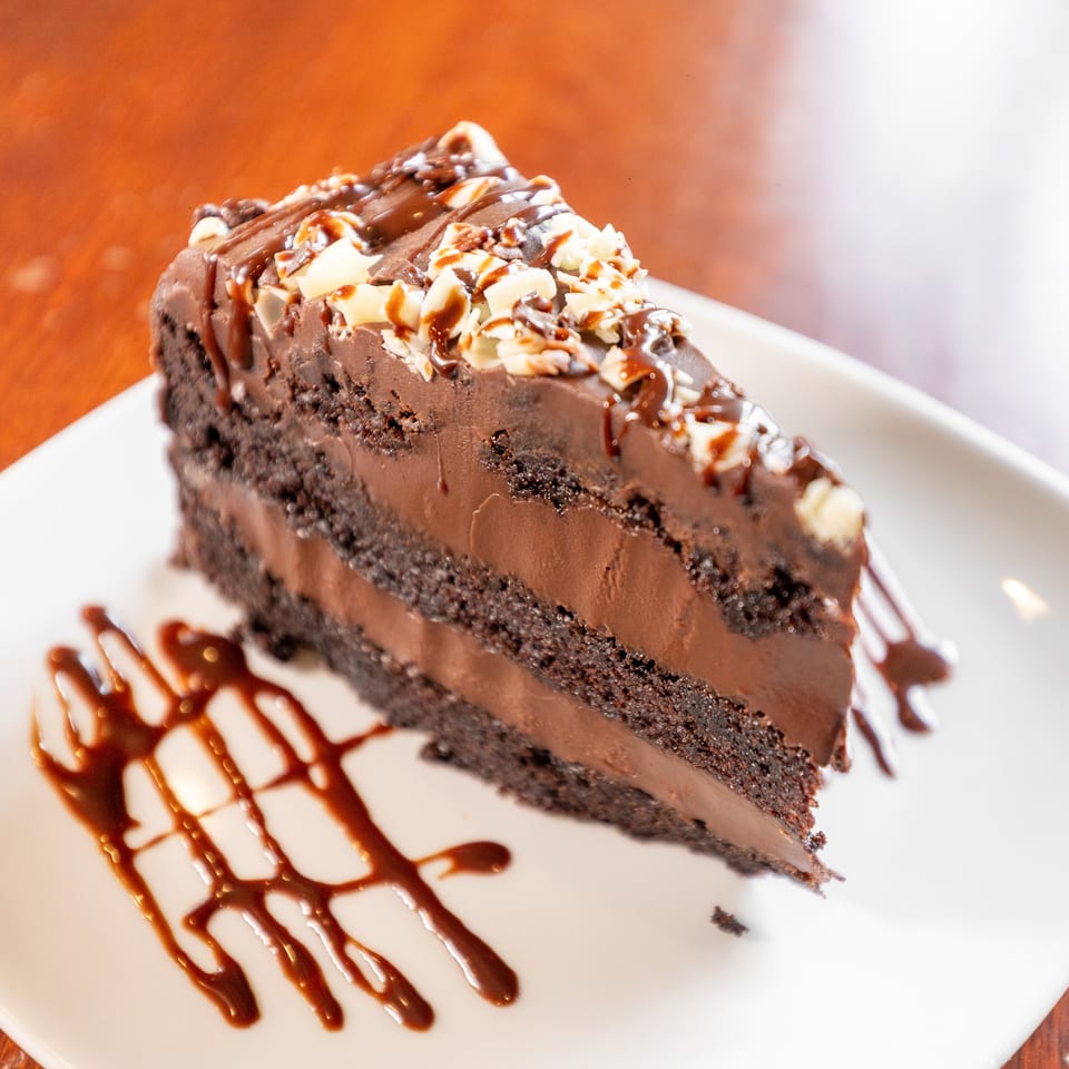 A slice of rich chocolate cake with chocolate frosting, drizzled with chocolate sauce and white chocolate shavings.