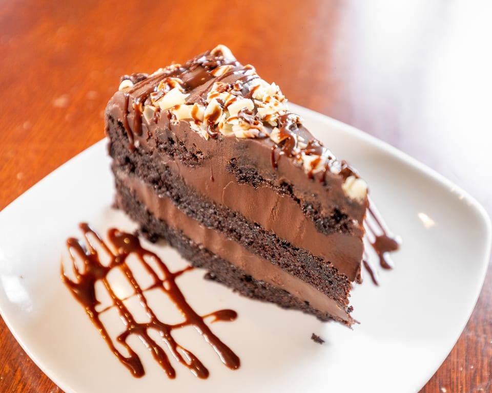 A slice of rich chocolate cake with chocolate frosting, drizzled with chocolate sauce and white chocolate shavings.