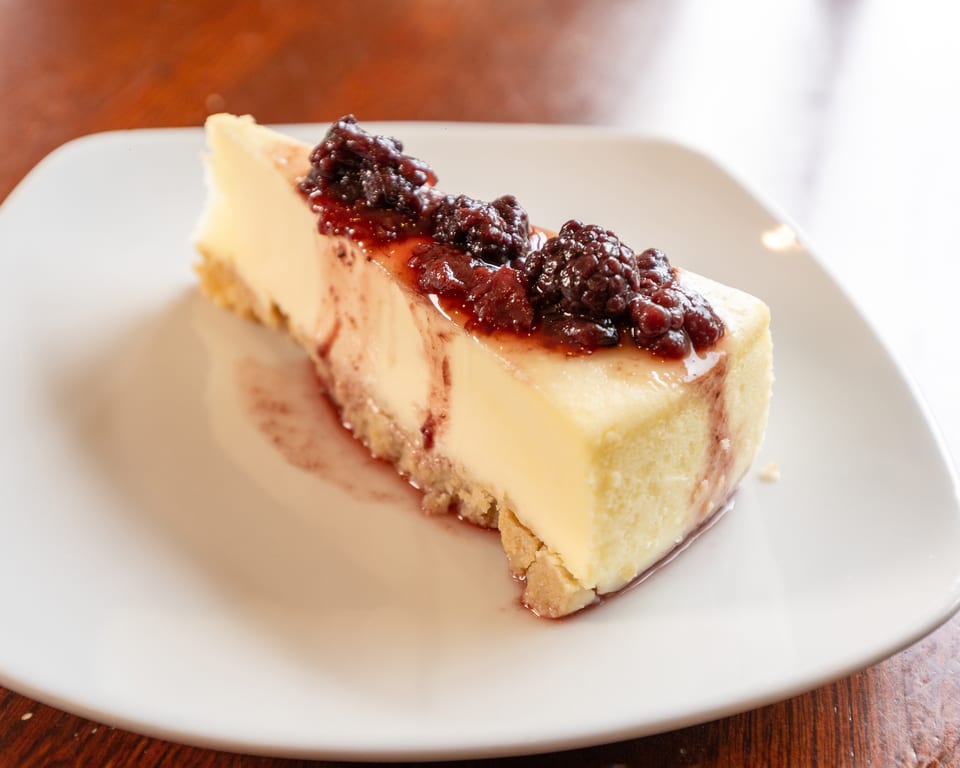 A slice of cheesecake with a graham cracker crust, topped with dark berry compote, on a white plate.